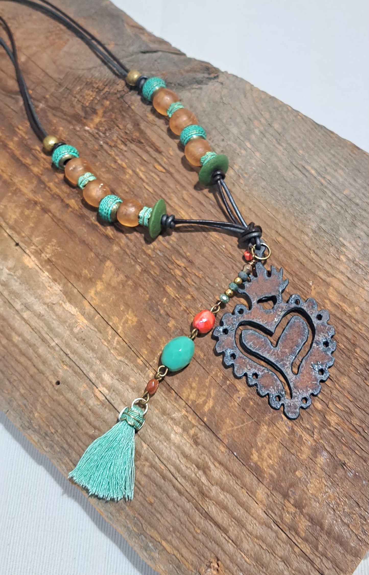 Necklace in grey and brown leather cord featuring a 2" iron heart charm decorated with blue and green beads and a sea green cotton tassel at its end.