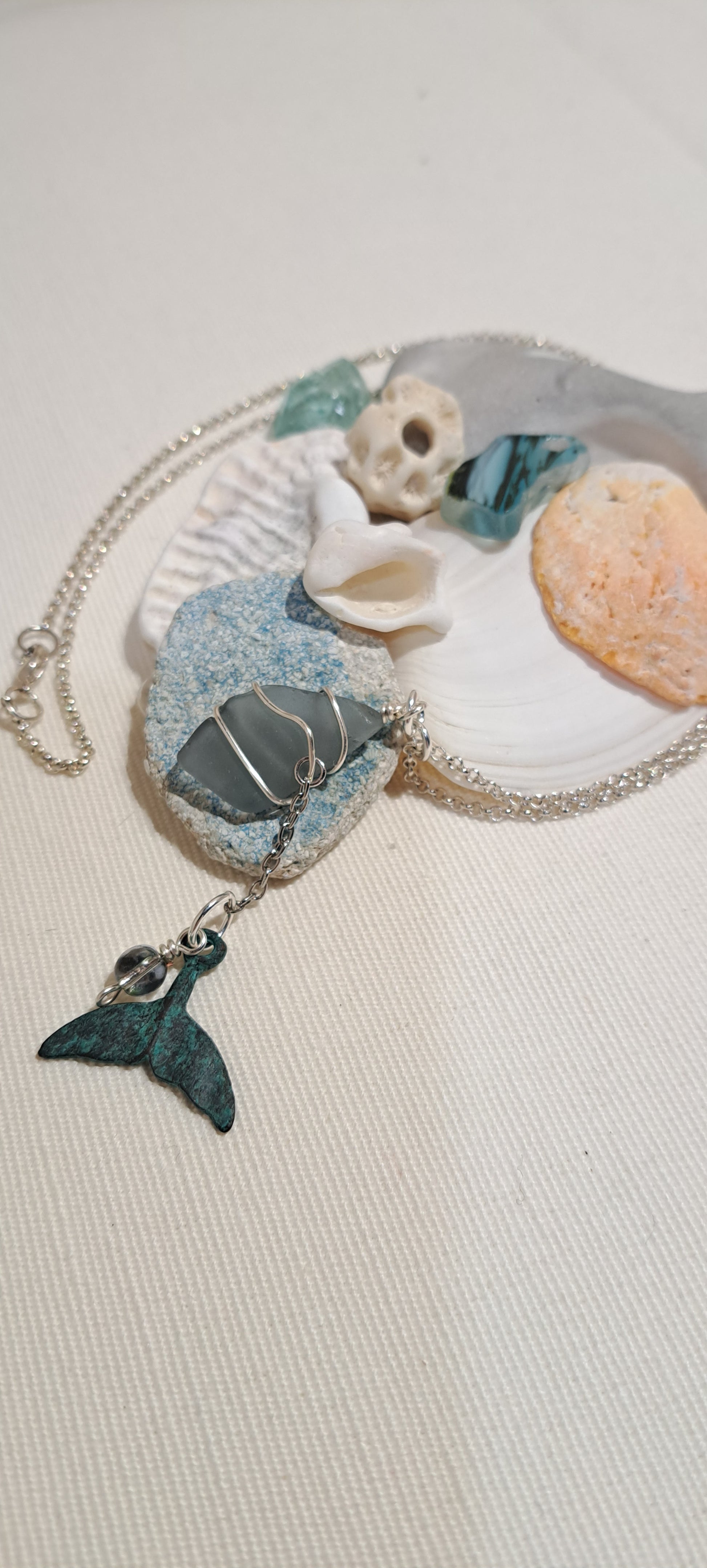 Necklace of stainless steel with a wire wrapped grey sea glass chip and a suspended green brass whale tail charm and tiny accent glass bead