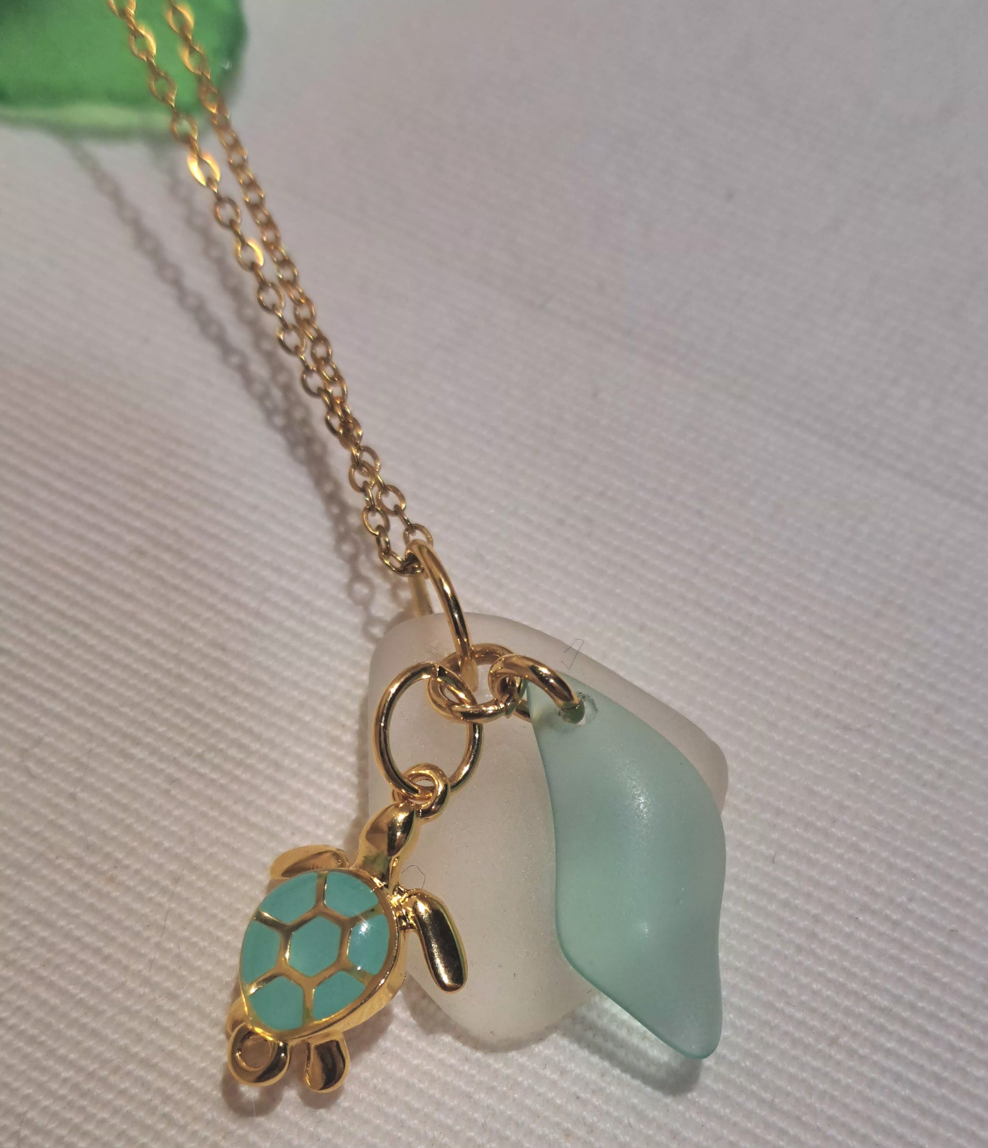 white sea glass piece hanging from a gold chain with a swirled bright green wave shaped glass and green and gold enamel turtle charm